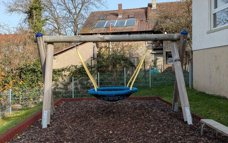 Blaue Nestschaukel an einem Holzgestell über einer Holzschnitzelfläche im Garten des Kindergartens